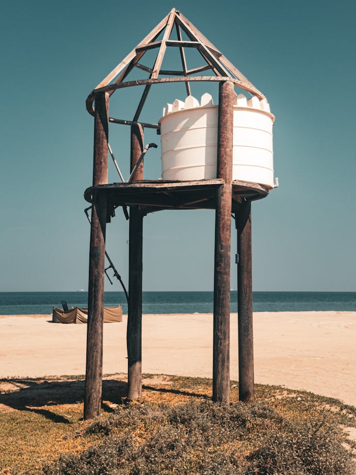 Minimalist wooden structure with water tank on a sandy beach under clear sky.